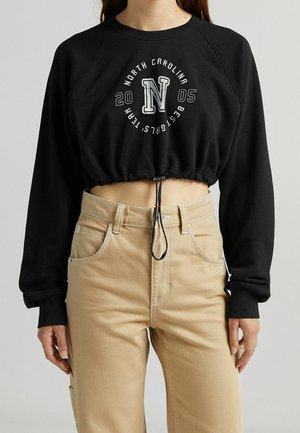 Personne portant un sweat-shirt court noir avec le texte "North Carolina 2005 Best Girls Team" et un pantalon beige taille haute.