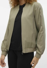 Oliwkowa bomberka z zamkiem z przodu, ściągaczami przy mankietach i gładką teksturą. Noszona na czarnej koszuli, z bocznymi kieszeniami.