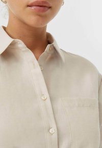 Close-up van een persoon die een lichtbeige overhemd met knopen en een kraag draagt, met een borstzak, waarbij de nek en het onderste deel van het gezicht zichtbaar zijn.