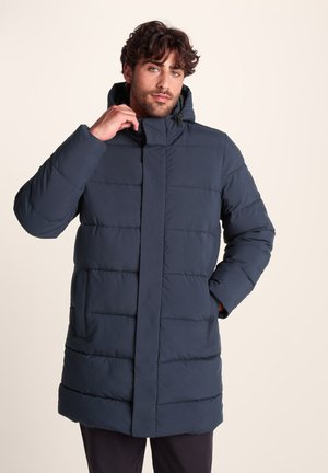 Man adjusting collar of navy blue quilted long puffer jacket with hood, standing against plain light background.