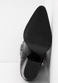 Botte noire en cuir à talon avec semelle pointue et talon empilé, vue par dessous sur fond blanc.