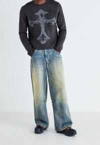 Grauer, flauschiger Pullover mit einem großen hellgrauen Kreuzdesign, kombiniert mit lockeren blauen Jeans aus Denim mit einem verblassten Farbverlauf und schwarzen Schuhen.