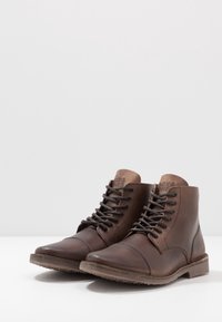 Pár hnědých kožených kotníkových bot na šněrování s kulatou špičkou a minimálním prošíváním na bílé ploše.