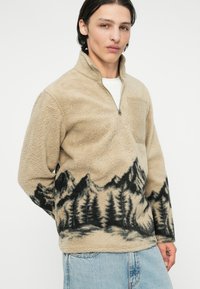 Beiger Fleece-Pullover mit einem halben Reißverschluss, der einen schwarzen Berg- und Waldlandschaftsdruck am Saum aufweist. Strukturierter Stoff.