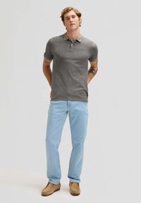 Hombre de pie con las manos detrás de la espalda, vistiendo un polo gris, jeans azul claro, zapatos marrones y tatuajes visibles en el brazo, sobre un fondo liso.