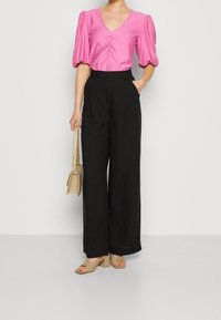 Růžová halenka s formovanými rukávy, výstřihem do V a nařasením; černé široké kalhoty; béžové sandály na blokovém podpatku a kabelka.