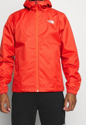 Klarrød jakke med lynlås og hætte, elastiske manchetter og The North Face-logo på brystet, båret med sorte bukser.