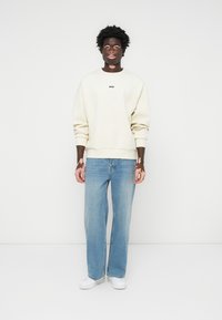 Sudadera de color crema claro con el logo "BOSS", combinada con unos vaqueros azules holgados y zapatillas blancas. El modelo se encuentra de pie frente a un fondo blanco liso.
