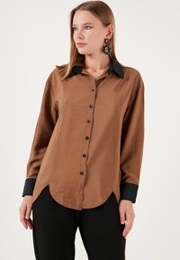 Brown satin button-up shirt with a black collar and cuffs, featuring a curved hem and black buttons, worn with black pants.