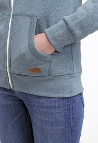 Felpa con zip, colore azzurro chiaro, dotata di tasche a marsupio, polsini e orlo a costine, e un'etichetta in pelle marrone sul davanti.