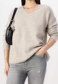 Pull en maille côtelée beige avec col en V et manches longues, associé à un jean gris légèrement déchiré et un petit sac bandoulière noir.