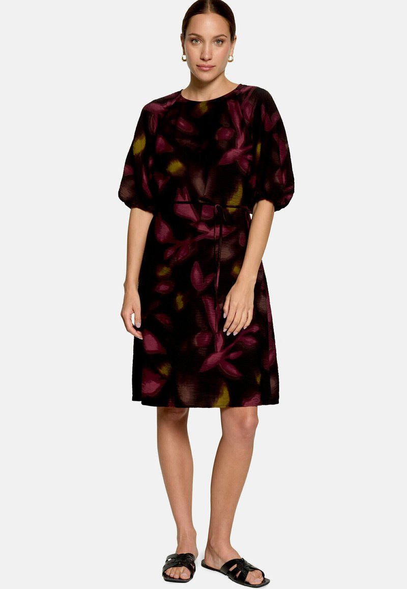 Femme aux cheveux attachés porte une robe noire et violette à fleurs, longueur genou, avec des manches bouffantes, une ceinture et des sandales plates noires.