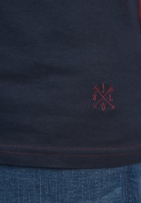 Navy cotton t-shirt with red stitching and embroidered graphic featuring crossed arrows at the hem. Smooth texture, casual design.
