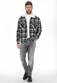 Chaqueta camisera de cuadros en blanco y negro con cuello de forro polar crema, dos bolsillos en el pecho, llevada sobre una camisa blanca, combinada con vaqueros grises y botas negras.