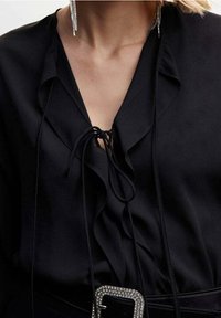 Blouse noire avec un col en V orné d'un détail à nouer, un devant à volants, et une ceinture structurée avec une boucle décorée de strass.