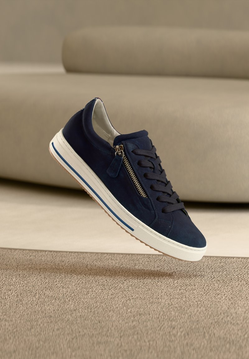 Σκούρο μπλε σουέντ αθλητικό παπούτσι με λευκή σόλα, σχέδιο με κορδόνια, πλαϊνό φερμουάρ και μπλε λωρίδα διακοσμητική λεπτομέρεια. Ανασηκωμένο πάνω από μια υφασμάτινη μπεζ επιφάνεια.