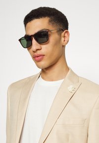 Ochelari de soare cu ramă tortoiseshell maro și lentile verzi, purtați cu o jachetă bej și un tricou alb. Include un ac de rever floral alb.