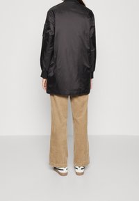 Černá saténová bomber bunda s dlouhými rukávy, žebrovanými manžetami a texturovanou nášivkou na levém rukávu, doplněná o široké kalhoty z kordu v béžové barvě.