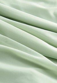 Plis de tissu vert clair doux avec une texture fine visible et un léger éclat, créant des motifs lisses et superposés.