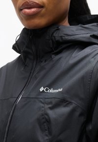 Κοντινό πλάνο ατόμου που φοράει μαύρο μπουφάν Columbia με κουκούλα και φερμουάρ, με το λογότυπο της μάρκας στο στήθος.