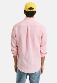 Lichtroze linnen shirt met geknoopte kraag, opgerolde mouwen en een plooi aan de achterkant, gedragen met blauwe jeans en een geel petje.