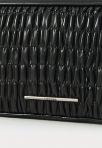 Juodas ketvirtasis odinis rankinės modelis su elegantišku sidabriniu "Calvin Klein" logotipu ir tekstūruotu raštu visame paviršiuje. Uždarymo detalė su užtrauktuku.