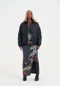 Černá kožená bunda přes modré šaty s grafikou ve stylu tetování; šaty mají vysoký rozparek a černé kozačky přes kolena s přezkami.