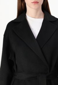 Abrigo de mezcla de lana negro con amplias solapas, cintura ceñida y mangas largas. Tejido suave y texturizado con detalles de costura mínimos y un ajuste relajado.
