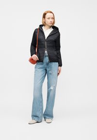 Γυναίκα που φοράει μαύρο καπιτονέ μπουφάν, λευκό πουκάμισο, τζιν παντελόνι με φαρδιές και σκισμένες μπατζάκια, μπεζ παπούτσια και κρατά μια μικρή κόκκινη τσάντα ώμου.