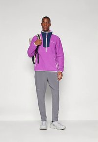 Herre lilla fleecepullover med marineblå detaljer, halv lynlås foran og to sidelommer, kombineret med grå taperede bukser og hvide sneakers.