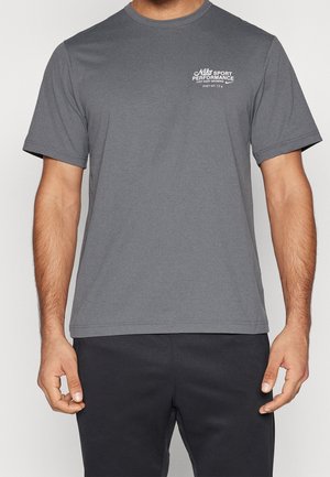 Homme portant un t-shirt Nike gris à manches courtes avec logo et texte blancs « "Nike Sport Performance" » sur la poitrine gauche, associé à un pantalon noir.