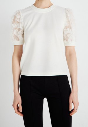 Biały top z krótkim rękawem i przezroczystymi kwiatowymi bufiastymi rękawkami w połączeniu z czarnymi dopasowanymi spodniami na stojącej osobie na jednolitym tle.