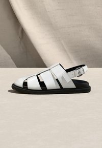 Λευκή δερμάτινη σαγιονάρα ψαρά με μαύρη σόλα και ρυθμιζόμενο λουρί με αγκράφα σε ουδέτερο υφασμάτινο φόντο.