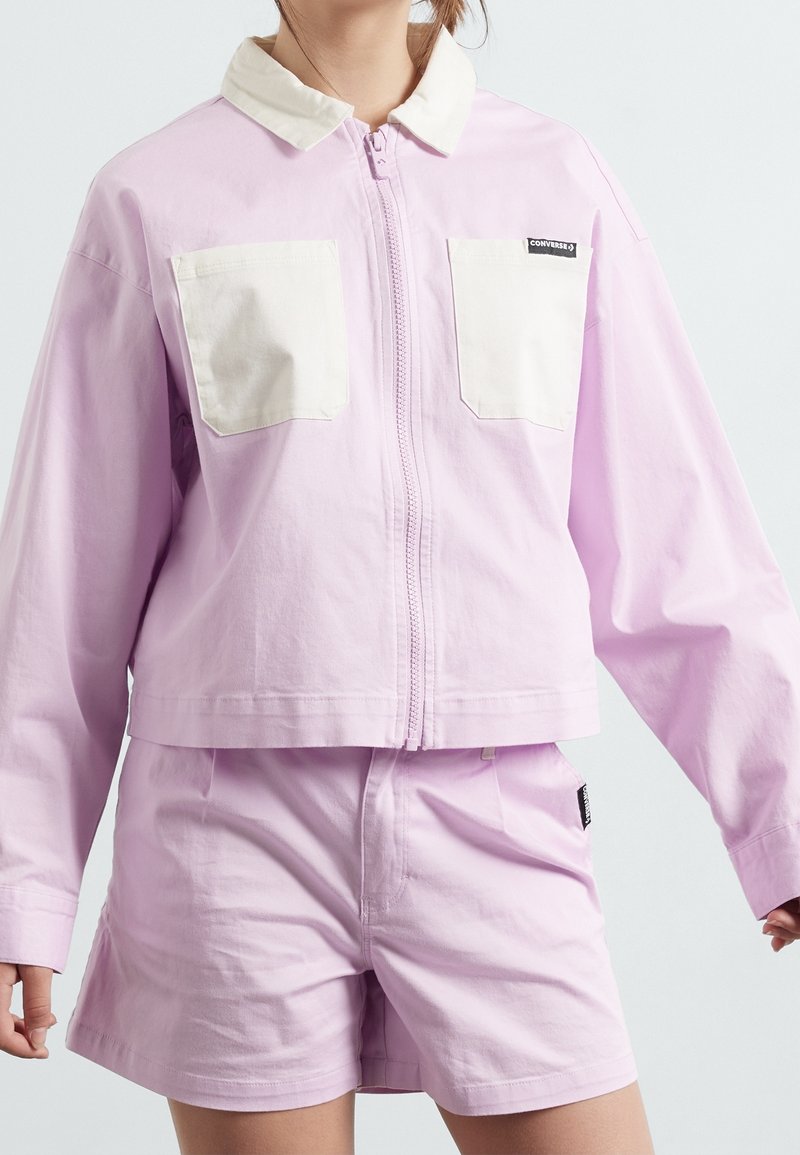 Personne portant une veste zippée violet clair avec des poches poitrine blanches et un short violet assorti, sur un fond uni.