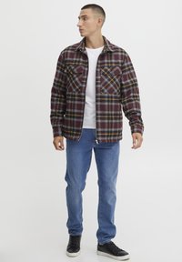 Geruite flanellen shirt in bordeaux, grijs en geel. Beschikt over twee borstzakken met kleppen. Gepaard met blauwe jeans en zwarte sneakers.