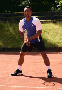 Atleet in een blauw en wit sporttop met zwarte shorts en zwarte tennisschoenen, die een tennisracket vasthoudt op een gravelbaan.