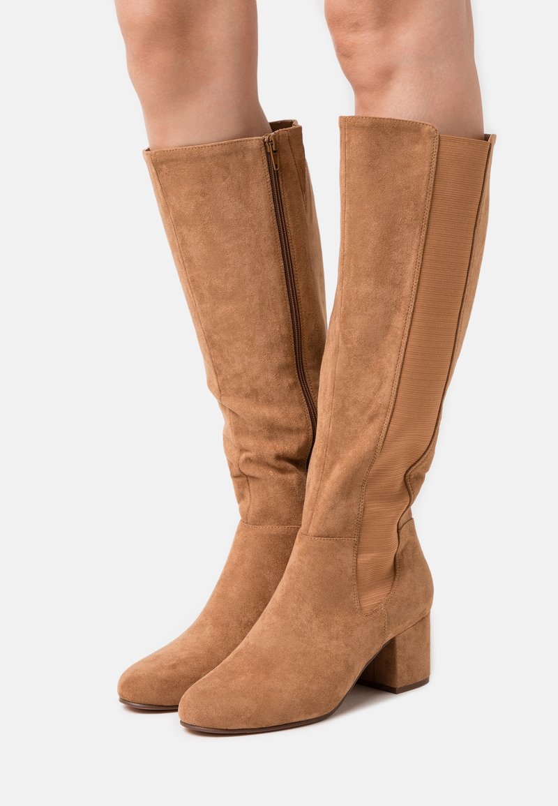 Bottes mi-mollet en daim beige avec talons blocs, fermeture éclair latérale sur une botte et panneau élastique sur l'autre, portées sur jambes nues.