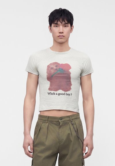 Νεαρός άνδρας φοράει κοντό ανοιχτό γκρι T-shirt με εικόνα σκύλου και το κείμενο "Who's a good boy?", συνδυασμένο με χακί παντελόνια.