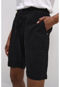 Personne portant un short noir au niveau des genoux avec des poches latérales, une main dans la poche, associé à un haut blanc rentré.