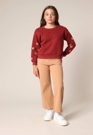Sweatshirt rouge avec des motifs en cœur dorés sur les manches, associé à un pantalon évasé marron clair et des baskets blanches. Tissu lisse, design décontracté.