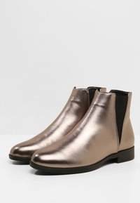 Metallinhohtoiset ruusukullanväriset nilkkurit, joissa sileä pinta, kaartuva kärki, joustavat sivupaneelit ja matala paksupohjakorko. Klassinen Chelsea-saappaan muotoilu.