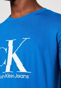 Modrá bavlněná tričko s kulatým výstřihem, na přední straně s velkým bílým logem "CK" a textem "Calvin Klein Jeans" na spodní části v hladké textuře.