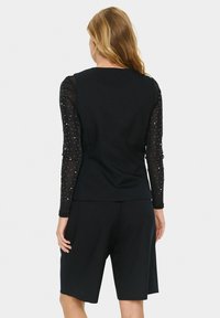 Schwarzes Outfit mit einem enganliegenden Oberteil mit durchsichtigen, verzierten Ärmeln und locker sitzenden Shorts. Glatte Textur und minimalistisches Design.