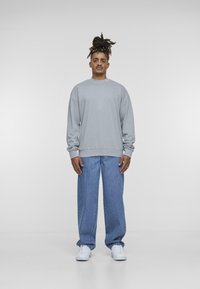 Grå hettegenser med lange ermer og ribbestrikkede mansjetter, kombinert med løse blå jeans og hvite joggesko, satt mot en ensfarget bakgrunn.