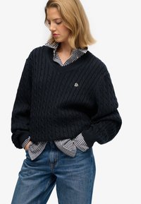 Woman with hands in pockets wearing navy cable knit sweater over black and white striped shirt and blue jeans against white background.