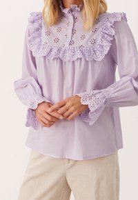 Blouse lavande claire avec bordure en dentelle ajourée sur le col et les manches. Dispose d'un devant boutonné et de poignets légèrement froncés.