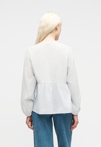 Γυναίκα με μακριά ξανθά μαλλιά φοράει μπλούζα με υφή σε ανοιχτό γαλάζιο και λευκό με μακριά μανίκια και μεσαίου τόνου μπλε τζιν, γυρισμένη με την πλάτη σε απλό φόντο.