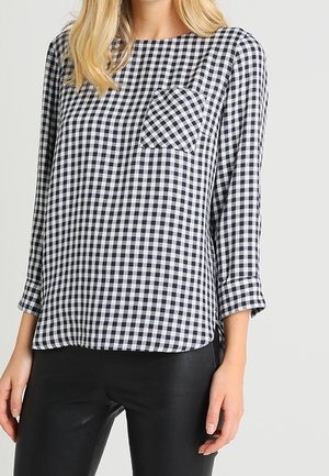 Kobieta nosząca czarno-białą, w kratę gingham, bluzkę z długim rękawem i przednią kieszenią, w połączeniu z czarnymi dopasowanymi spodniami.