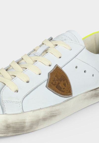 Baskets blanches avec lacets crème, écusson marron en forme de blason sur le côté, semelle légèrement sale et détail jaune fluo visible sur la partie intérieure du talon.