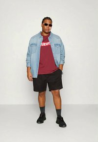 Denim button-up shirt over een kastanjebruine Levi's t-shirt en zwarte shorts. Zwarte sneakers en grijze sokken maken de outfit compleet.